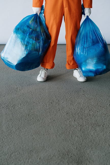 A person dressed in orange trousers and white sneakers is standing indoors on a grey carpeted floor, holding two large blue plastic refuse bags filled with bulky waste. The bags appear to contain soft materials or household rubbish, with some items possibly wrapped in plastic or fabric for disposal. The individual is holding one bag in each hand, with arms slightly bent, positioned near the hips. Behind them, a plain white wall can be seen, suggesting an interior setting such as a home or storage area. This scene relates to the process of home clearance or furniture transport, typical of a house removals service like those offered by Man With a Van Merton, who operate within the context of professional packing, loading, and transportation logistics involved in moves and waste collection in Merton.