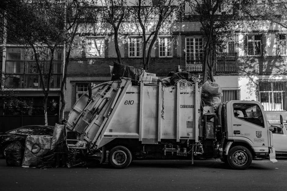A black-and-white photograph showing a rubbish collection truck parked on a residential street in Merton during daytime. The truck is loaded with various waste objects, including large black garbage bags, cardboard boxes, and household items, which are positioned in the back compartment. The vehicle appears to be in the process of loading or unloading waste, with some items secured with straps or wrapped in plastic. The setting includes an apartment building in the background with multiple windows, some with metal bars, and leafless trees lining the street. The truck's side features a company name and logo, representing Man With a Van Merton, a removals service provider. The scene illustrates typical home clearance or waste removal activities associated with household moves or renovation projects, emphasizing logistical aspects of furniture transport and packing materials connected to house removals and council waste collection rules in Merton.