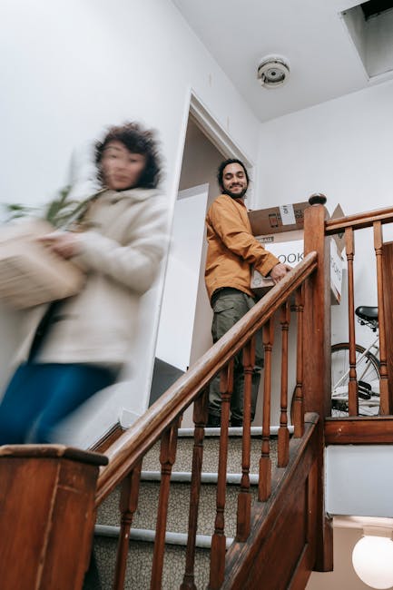 Inside a residential property, a man with a beard, wearing a brown jacket, is standing on a staircase landing holding a cardboard box, ready for home relocation. A woman in a beige coat is seen in motion carrying another box, appearing slightly blurred due to movement. The staircase features wooden bannisters and carpeted steps, and the background includes a doorway leading into a room with white walls and a ceiling-mounted smoke detector. On the staircase, there are additional packed cardboard boxes, some sealed with packing tape, indicating the process of packing and moving. Nearby, a bicycle is visible, and the environment is well-lit with natural light from an unseen window. This scene is part of a furniture transport and packing process facilitated by Man With a Van Merton, specialising in removals, supporting efficient home relocation in Colliers Wood with estate stair and lift access.
