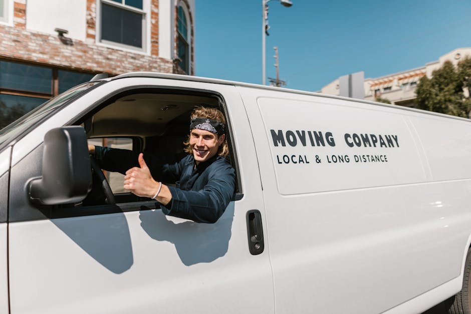 A smiling young man wearing a black bandana and dark clothing is sitting in the driver's seat of a white moving van, giving a thumbs-up gesture. The van has the text 'MOVING COMPANY LOCAL & LONG DISTA