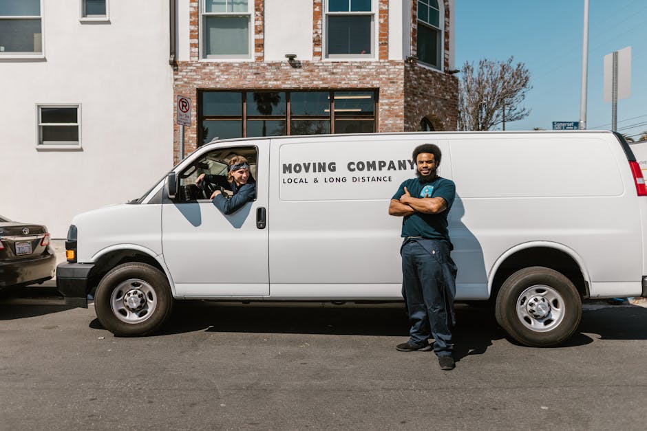 A smiling young man wearing a black bandana and dark clothing is sitting in the driver's seat of a white moving van, giving a thumbs-up gesture. The van has the text 'MOVING COMPANY LOCAL & LONG DISTA