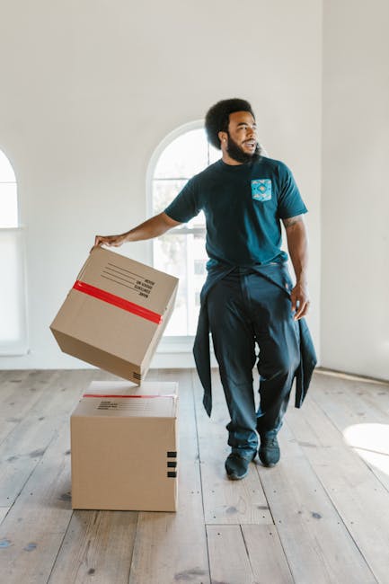 A male mover with dark skin and an afro hairstyle is inside a bright, airy room with white walls and wooden flooring. He is dressed in a blue t-shirt with a light blue logo on the chest, dark trousers, and is holding a cardboard box wrapped with red tape in his right hand, preparing to place it on the floor. There are two cardboard boxes stacked in front of him, one of which he is handling, indicating the packing and loading process involved in home relocation. Natural daylight streams through a large arched window behind him, illuminating the scene. The environment appears to be in the midst of a household move, with minimal furnishings visible. The presence of these moving boxes and the man's activity reflects the logistics and physical effort associated with local removals and furniture transport, which Man With a Van Merton provides as part of their house removal services.