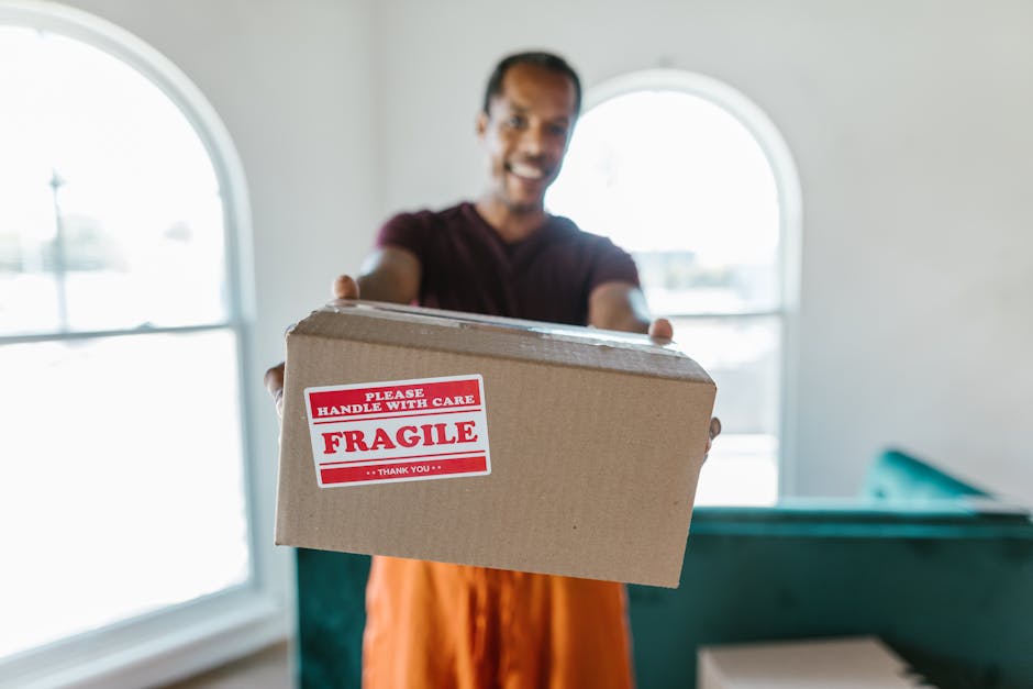 A man wearing a maroon t-shirt and orange trousers is standing inside a well-lit room with white walls and two arched windows in the background, holding out a medium-sized cardboard box towards the camera. The box has a red and white label that reads 'Please Handle with Care, Fragile, Thank You,' indicating it contains delicate items. The scene suggests the process of home relocation or packing and moving, with the man preparing to load or transport the box. The room appears to be part of a house being prepared for a move, and the focus on the box and the man's gesture highlights careful handling during furniture transport or packing activities. Occasionally, [COMPANY_NAME] might be involved in such moving and removals services, which often include handling fragile items like these during residential moves.