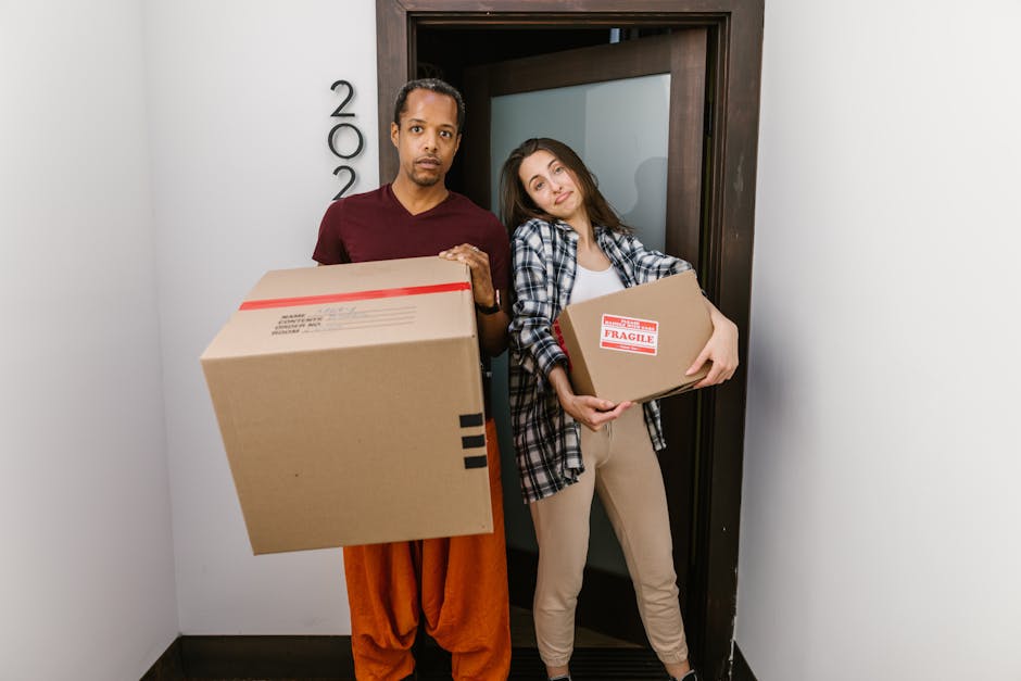 A male mover with dark skin and an afro hairstyle is inside a bright, airy room with white walls and wooden flooring. He is dressed in a blue t-shirt with a light blue logo on the chest, dark trousers, and is holding a cardboard box wrapped with red tape in his right hand, preparing to place it on the floor. There are two cardboard boxes stacked in front of him, one of which he is handling, indicating the packing and loading process involved in home relocation. Natural daylight streams through a large arched window behind him, illuminating the scene. The environment appears to be in the midst of a household move, with minimal furnishings visible. The presence of these moving boxes and the man's activity reflects the logistics and physical effort associated with local removals and furniture transport, which Man With a Van Merton provides as part of their house removal services.