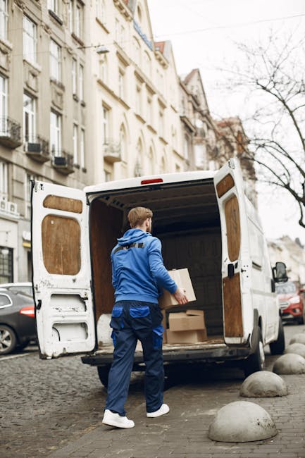 A man in a blue jacket and dark trousers loads several cardboard boxes into the open rear of a white moving van parked on a cobblestone street. The van's interior shows wooden panels and the back doors are open, revealing the interior space designated for furniture and packing materials. The street is lined with multi-storey residential buildings featuring classic architectural details, and there are parked cars and large, rounded concrete bollards along the pavement. The scene indicates an ongoing home relocation or furniture transport process, with the man preparing for or in the middle of a house removal. This image exemplifies packing, loading, and transport activities central to professional removals services, with the focus on careful handling of boxes and efficient use of vehicle space, consistent with services offered by companies like Man With a Van Merton.