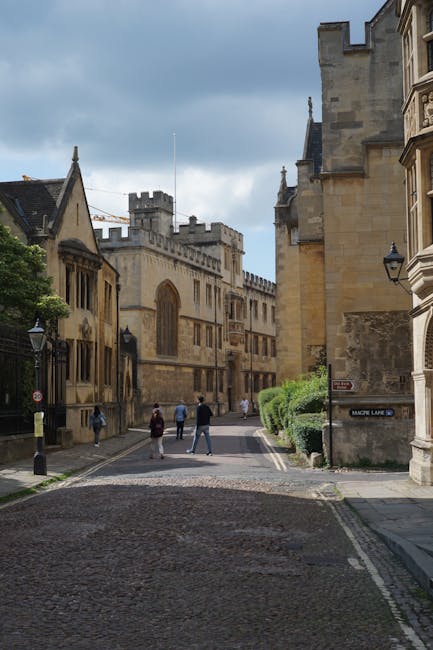 A city street scene with historic stone buildings lining both sides, including a large church or castle-like structure with pointed arches and battlements in the background. The cobblestone road is narrow and slightly curved, with a small curb and street lamps along the pavement. Several pedestrians are walking along the sidewalk, some in pairs and others alone, dressed in casual clothing. On the right side, a building has a small sign reading 'Magpie Lane' attached to the wall, and neatly trimmed shrubs grow adjacent to it. The sky above is partly cloudy with patches of blue visible, suggesting daytime. This environment reflects an urban setting suitable for a house or furniture removal service, highlighting the area's architecture and transportation possibilities for relocations, in line with the services offered by Man With a Van Merton.