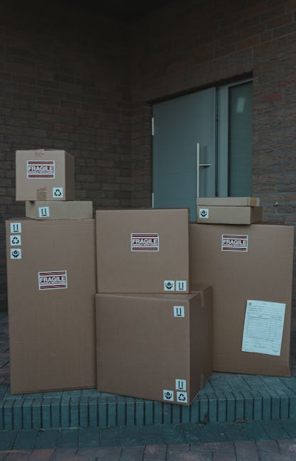 A man in a blue jacket and dark trousers loads several cardboard boxes into the open rear of a white moving van parked on a cobblestone street. The van's interior shows wooden panels and the back doors are open, revealing the interior space designated for furniture and packing materials. The street is lined with multi-storey residential buildings featuring classic architectural details, and there are parked cars and large, rounded concrete bollards along the pavement. The scene indicates an ongoing home relocation or furniture transport process, with the man preparing for or in the middle of a house removal. This image exemplifies packing, loading, and transport activities central to professional removals services, with the focus on careful handling of boxes and efficient use of vehicle space, consistent with services offered by companies like Man With a Van Merton.
