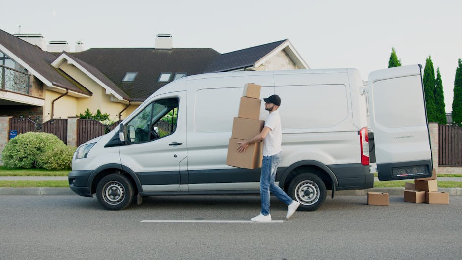 A man wearing a white t-shirt, blue jeans, white sneakers, and a cap is carrying three stacked cardboard boxes, with additional boxes placed on the pavement nearby, in front of a white commercial van parked on a residential street. The van is part of a home relocation and furniture transport process, with the rear doors open, indicating loading or unloading of items. The background features a modern house with a sloped roof, multiple windows, and a garden with well-maintained shrubs and tall trees, under bright daytime lighting. The scene depicts a professional removals operation, likely supported by a moving service such as Man With a Van Merton, focused on packing and moving tasks, including furniture and box transportation, to accommodate house moves or property transfers in a suburban neighbourhood.