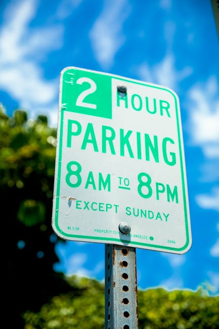 A green and white parking restriction sign attached to a metal pole, indicating a 2-hour parking limit from 8 a.m. to 8 p.m. daily, excluding Sundays, positioned outdoors against a backdrop of a blue sky with scattered clouds and blurred green foliage. The sign is part of urban traffic management, relevant for movers and removal companies like Man With a Van Merton when planning parking permits for home relocation or furniture transport, ensuring compliance with local parking rules during loading and unloading operations.
