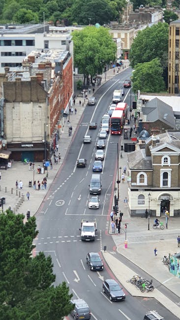 An aerial view of a busy urban street in Merton, showing a line of vehicles including cars and a double-decker bus moving along the road. On the sides of the street, there are various buildings such as commercial shops, residential houses, and a small church with a grey roof. Sidewalks are occupied by pedestrians, some walking and others waiting to cross the road. There are trees lining the street, providing greenery amidst the urban environment. The scene captures the typical activity during daytime, with clear weather and good visibility, illustrating the kind of setting in which a professional house removal service like Man With a Van Merton might operate to facilitate efficient furniture transport and home relocation tasks, including loading and unloading process within a local community.