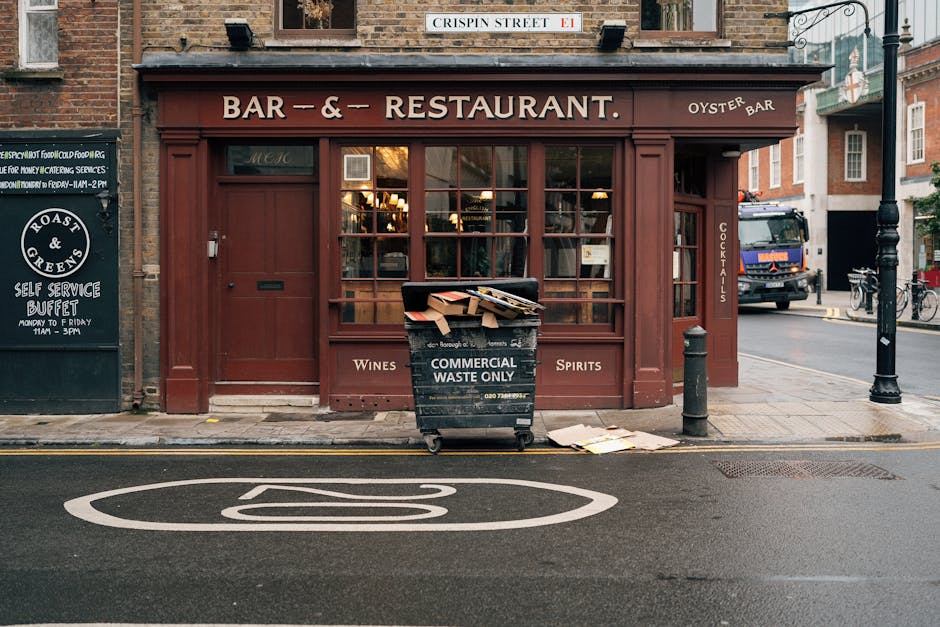 A street view showing the front of a closed bar and restaurant with a deep red facade, large glass windows, and a wooden door at the center. In front of the establishment, a black wheeled waste bin marked 'Commercial Waste Only' is filled with flattened cardboard boxes and paper, with some trash spilling onto the sidewalk. The bin is positioned next to a grey bollard and a small pile of flattened cardboard on the pavement. To the left, a blackboard sign advertises a self-service buffet, while on the right, a black street lamp and a blue delivery truck are visible further down the road. The scene is illuminated by natural light, suggesting early evening, with the surrounding environment including other brick buildings and street elements consistent with Merton, London. The image captures a moment of logistical activity related to household or business packing and waste disposal, which aligns with professional removals and home relocation services advocated by Man With a Van Merton.