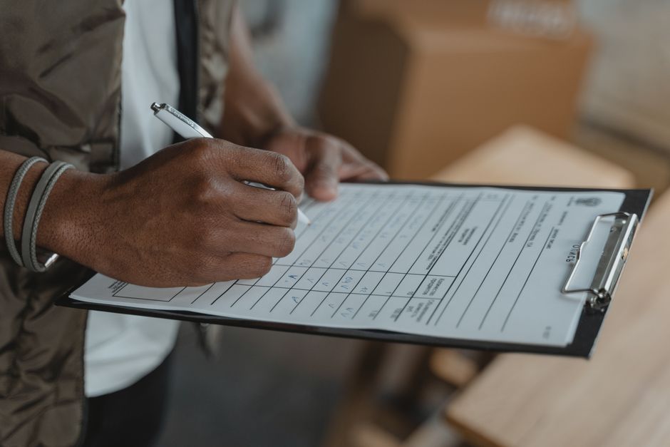 Close-up of a person with dark skin, wearing a gray wristband, holding a silver pen and filling out a checklist on a clipboard with a printed form. The form contains rows and columns for recording details related to house removals, such as items, quantities, and packing statuses. The individual is inside a room with cardboard boxes and packaging materials visible in the background, indicating a packing or home relocation process. The scene emphasizes careful planning and organization during a furniture transport or moving operation, with the person’s hand and part of their arm in focus, while the background is slightly blurred. This image illustrates the detailed documentation involved in professional removals by [COMPANY_NAME], supporting services like packing, loading, and transportation of household items during a house move.