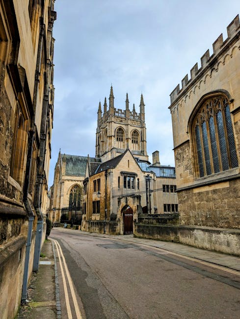 The image shows a narrow cobblestone street flanked by historic stone buildings with large gothic-style windows and decorative architectural details, leading towards a prominent medieval church with a tall, ornate tower topped by pinnacles, situated under a partly cloudy sky. The scene captures a quiet urban area, typical of a European city center, with no visible vehicles or people. Inside a property, near the entrance, there are several cardboard boxes sealed with packing tape, some covered with plastic wrap, alongside a few pieces of wooden furniture including a chair and a small table, as well as a fabric-covered item, possibly a sofa or mattress. A few moving blankets are spread on the floor, and a trolley with wheels is positioned nearby, suggesting recent or ongoing furniture transport as part of a house or flat removal process. The scene appears to be inside or at the entrance of a residential property, with the street and church visible in the background, supporting the context of house removals or furniture transport services provided by Man With a Van Merton for purposes like flat clearances or home relocations.