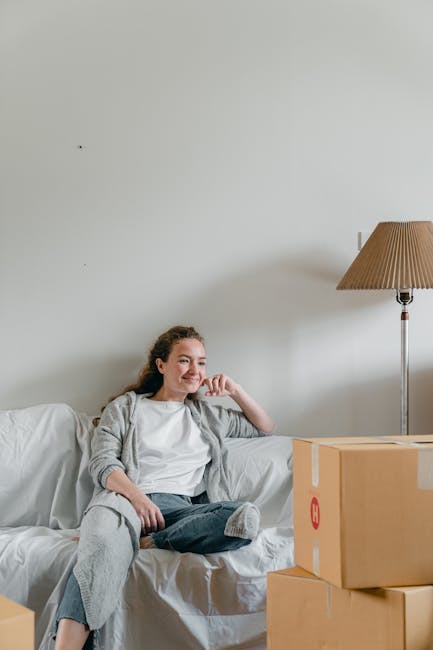 The image shows a young woman with long, curly hair sitting on a white sofa inside a room with plain white walls. She is smiling and appears relaxed, wearing a light grey cardigan over a white top and dark jeans. Beside her on the sofa, there are several cardboard boxes, some sealed with packing tape, indicating the process of packing for a move. To her right, a tall floor lamp with a conical beige shade illuminates the room. The scene suggests a home environment during a house clearance or packing process as part of a furniture transport or relocation service. This setting is consistent with the services offered by Man With a Van Merton, specifically focused on house removals and moving logistics, emphasizing packing and moving preparations.