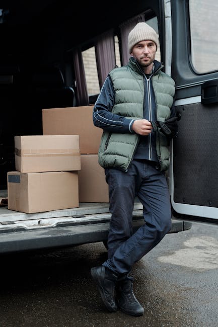A man dressed in a beige knit beanie, a green puffer vest over a navy blue jacket, dark trousers, and black boots is standing beside an open van on a paved street during daylight hours. Inside the van's loading area, several cardboard boxes of various sizes are visible, some stacked on top of each other. The man is holding a mobile phone in his right hand and appears to be in the process of organizing or preparing for a home relocation, with a focus on furniture transport and packing materials as part of a professional removals service. The van's interior is partially visible, showing the vehicle's dark seat upholstery and the side panels, suggesting a typical moving vehicle used by companies like Man With a Van Merton for residential moves and moving logistics. The scene reflects an organized loading or unloading process, supporting the context of house removals or furniture transport.