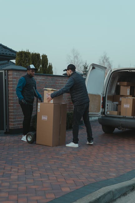 Two men are engaged in a home relocation process outside a residential property, with a van parked on a brick driveway. One man, wearing a black cap, black vest, and dark trousers, is lifting a cardboard box with both hands, preparing to load it into the open rear door of the van. The other man, dressed in a dark jacket and trousers, stands on a small hand trolley or dolly, waiting to assist. Several neatly packed cardboard boxes of varying sizes are visible, with labels indicating they are ready for transport. The van's interior appears to contain more boxes and packaging materials, suggesting a furniture transport and packing process as part of the moving service provided by Man With a Van Merton. The background includes a brick wall, some trees, and overcast sky, indicating a typical residential setting for domestic removals.