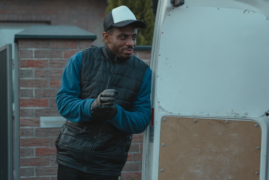 A man dressed in a beige knit beanie, a green puffer vest over a navy blue jacket, dark trousers, and black boots is standing beside an open van on a paved street during daylight hours. Inside the van's loading area, several cardboard boxes of various sizes are visible, some stacked on top of each other. The man is holding a mobile phone in his right hand and appears to be in the process of organizing or preparing for a home relocation, with a focus on furniture transport and packing materials as part of a professional removals service. The van's interior is partially visible, showing the vehicle's dark seat upholstery and the side panels, suggesting a typical moving vehicle used by companies like Man With a Van Merton for residential moves and moving logistics. The scene reflects an organized loading or unloading process, supporting the context of house removals or furniture transport.